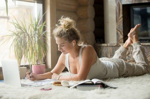 smiling young woman lying on white carpet studying with books and laptop in modern living room