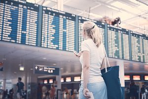 Young blonde woman with handbag, phone and tickets in her hands checking flight timetable in airport
